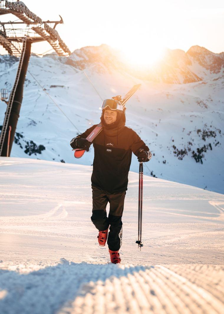 Smiling skier walking uphill at sunrise with skis on shoulder in alpine ski resort